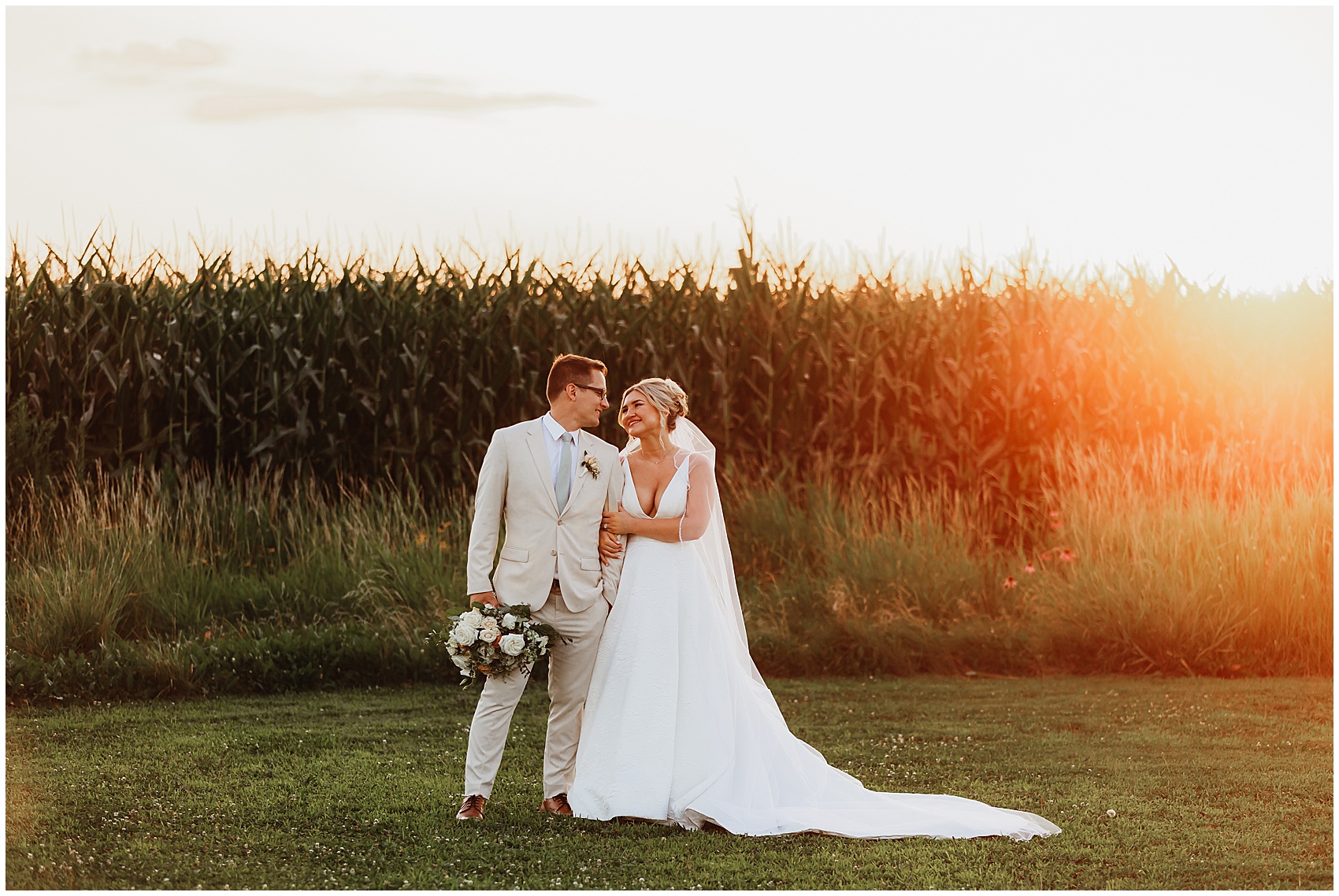 A bride in a long white silk dress holds onto the arm of her groom in a tan suit at sunset by a cornfield
