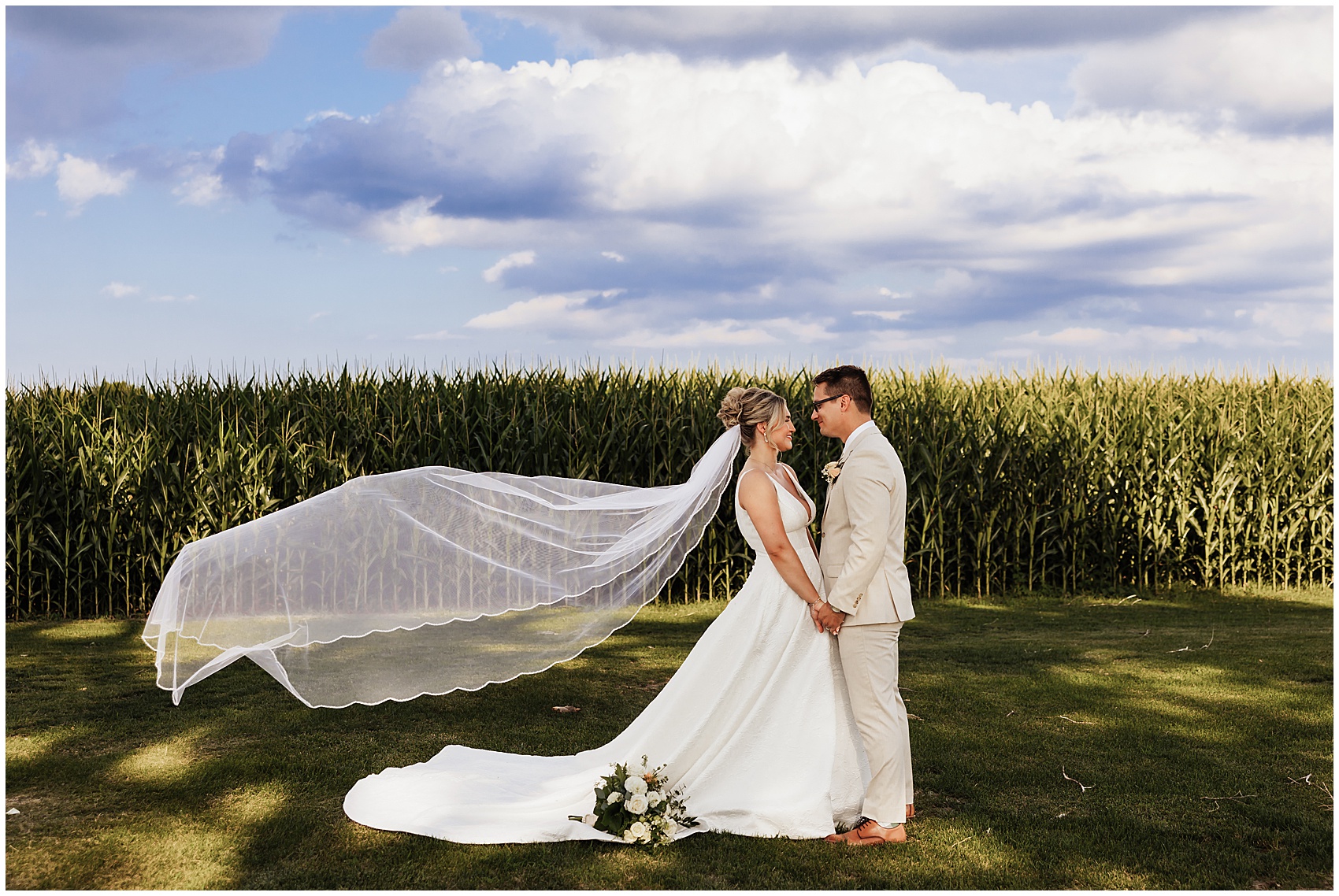 Newlyweds hold hands and stare into each other's eyes while the bride's veil flows behind her on the edge of a cornfield at a sable creek homestead wedding