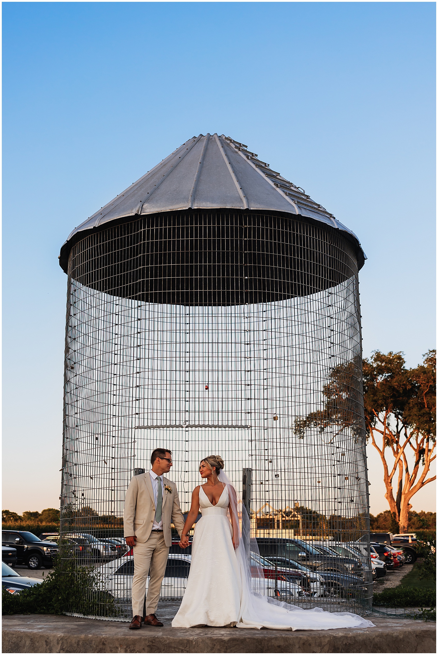 Newlyweds hold hands and gaze at each other while standing in front of a metal wire silo covered with padlocks sable creek homestead