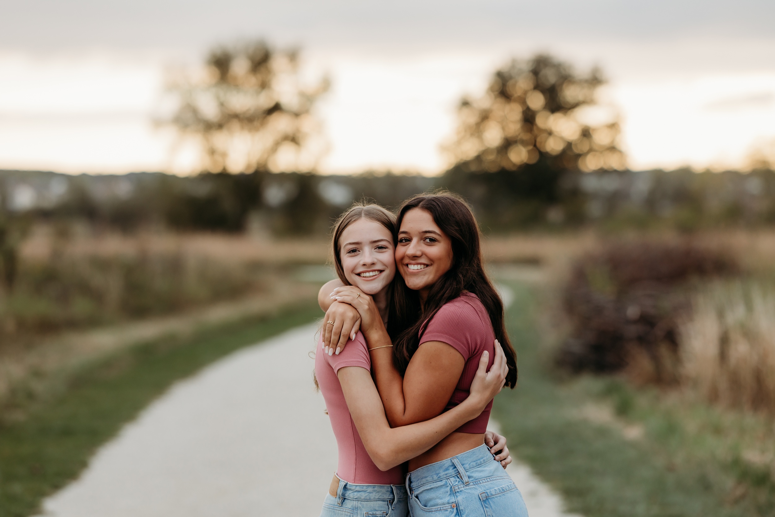 Best friends in matching pink tops and jeans hug while in a park at sunset